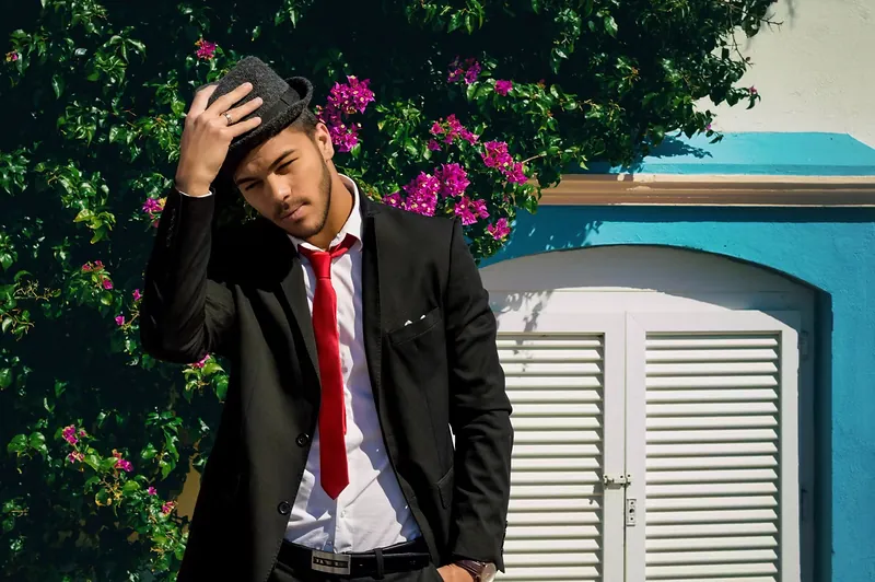 Modelo masculino con traje negro y corbata roja ante una fachada azul con buganvilla