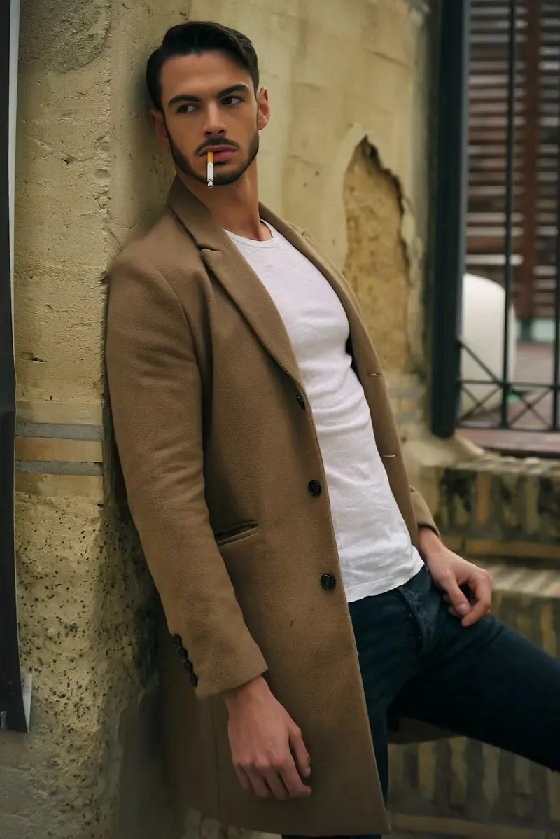 Modelo masculino con abrigo camel apoyado en un muro de piedra