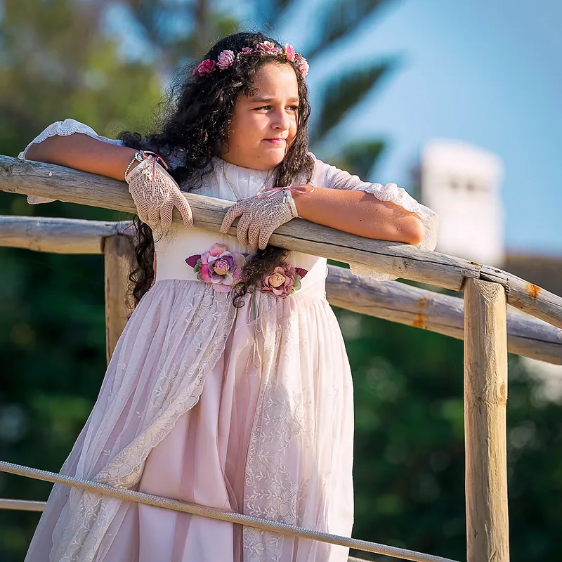 Niña de comunión con vestido rosa apoyada en una barandilla de madera