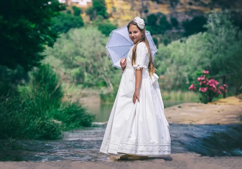 Niña de comunión con sombrilla blanca junto al río, entre la vegetación