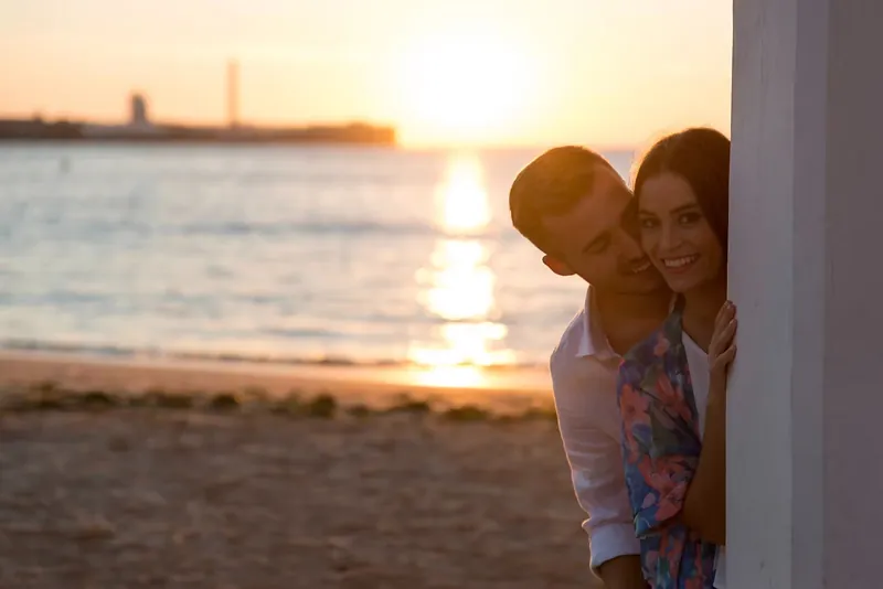 Pareja asomada tras una columna con la playa y el atardecer de fondo