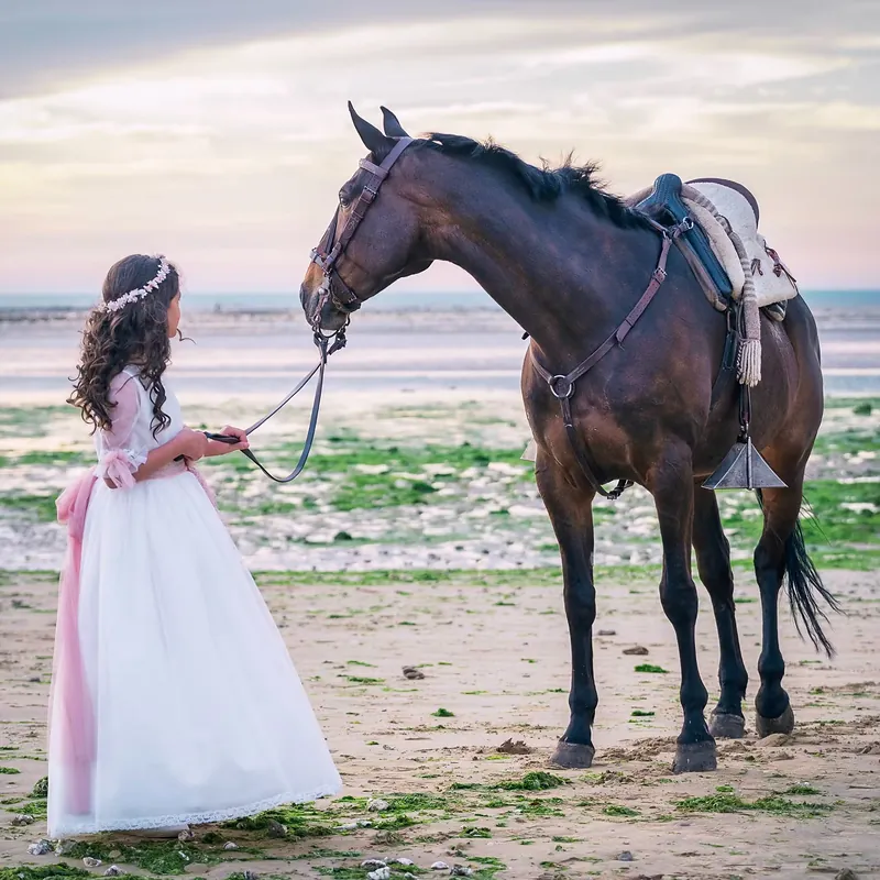 Niña de comunión llevando un caballo del ronzal por la playa al atardecer